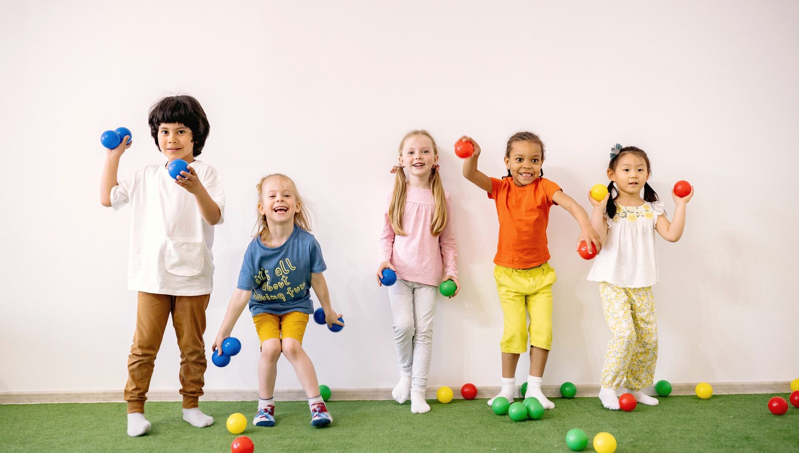 Five children hold colorful balls while sitting on green turf