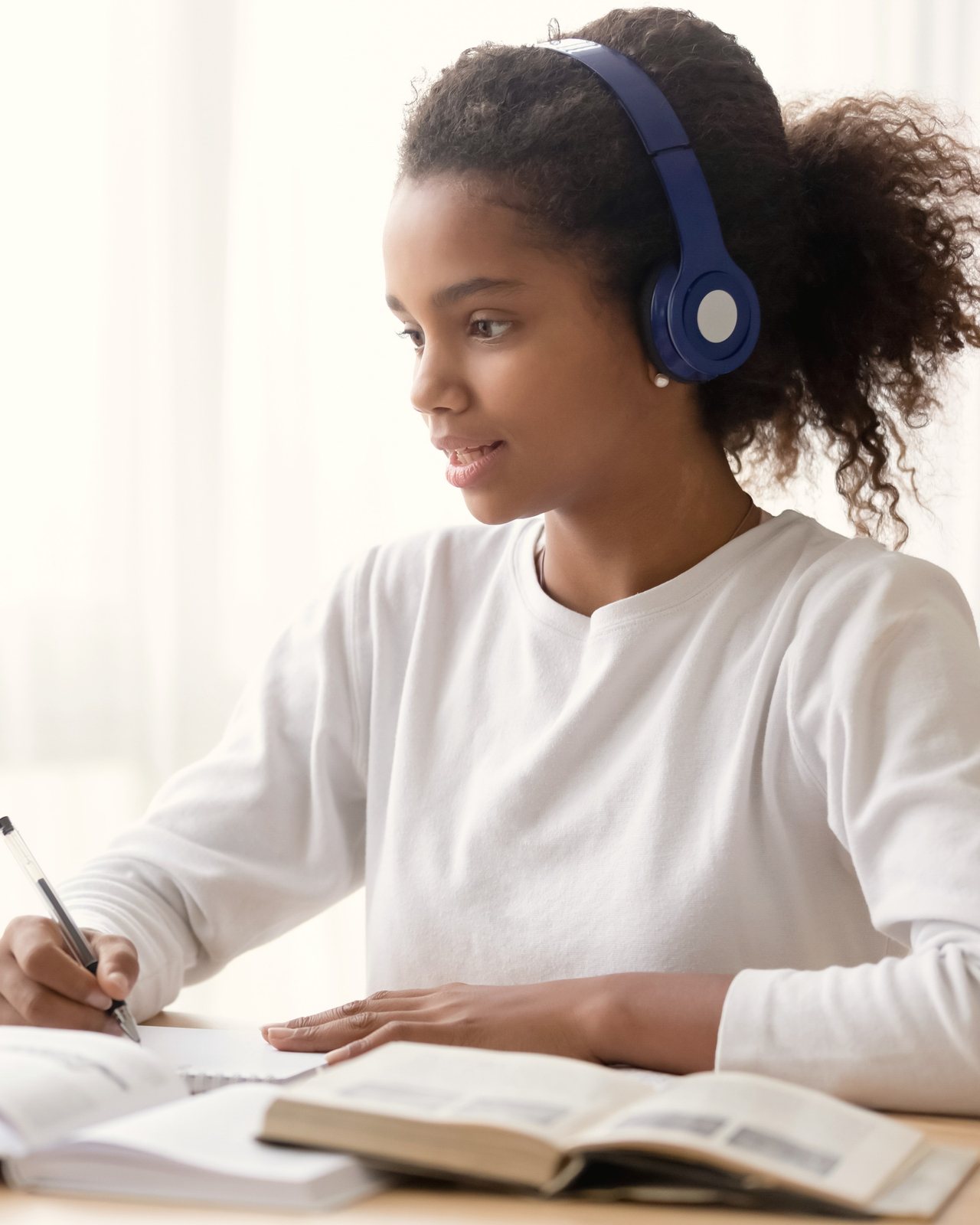 A teenage girl wearing headphones writes in a notebook next to her laptop