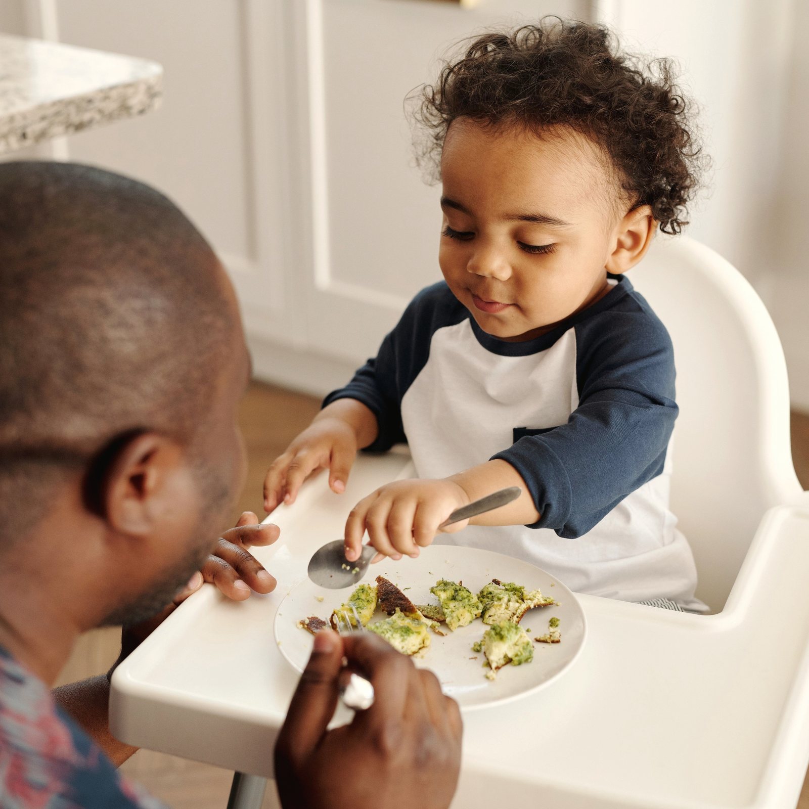 A dad spoon-feeds his smiling toddler in a high chair