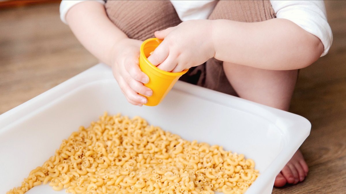 Toddler scooping dry pasta with a small yellow cup from a play bin, a simple fine motor activity at home