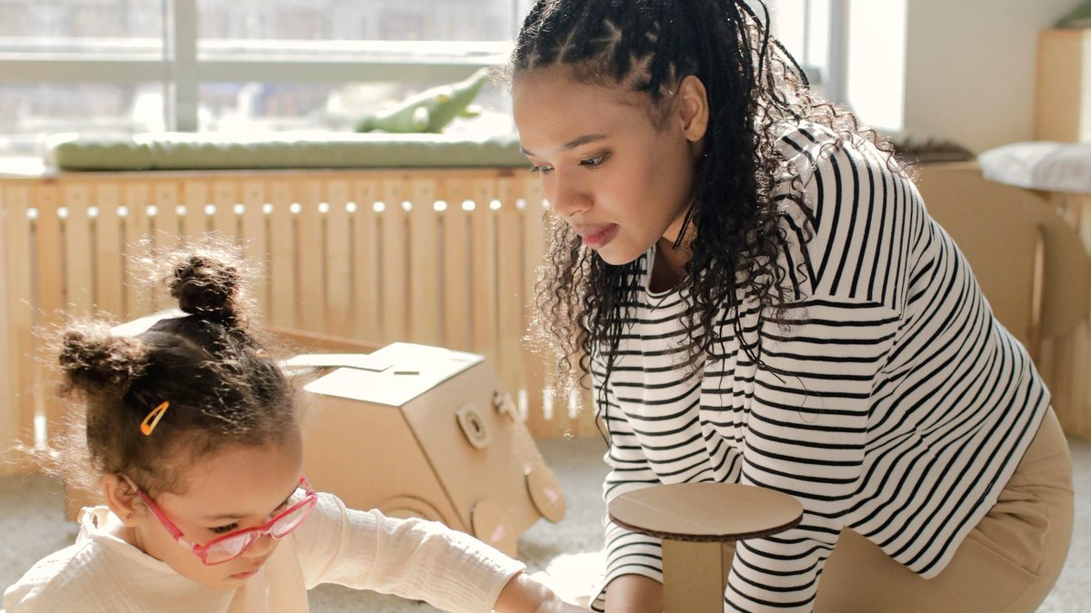 Woman and young girl in pink glasses building a cardboard car together on a playroom floor